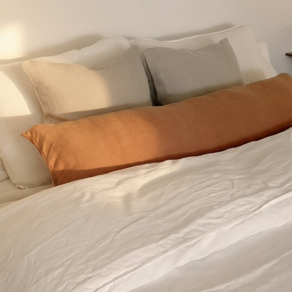 Bed with white duvet cover and beige and terracotta pillows against a white wall.