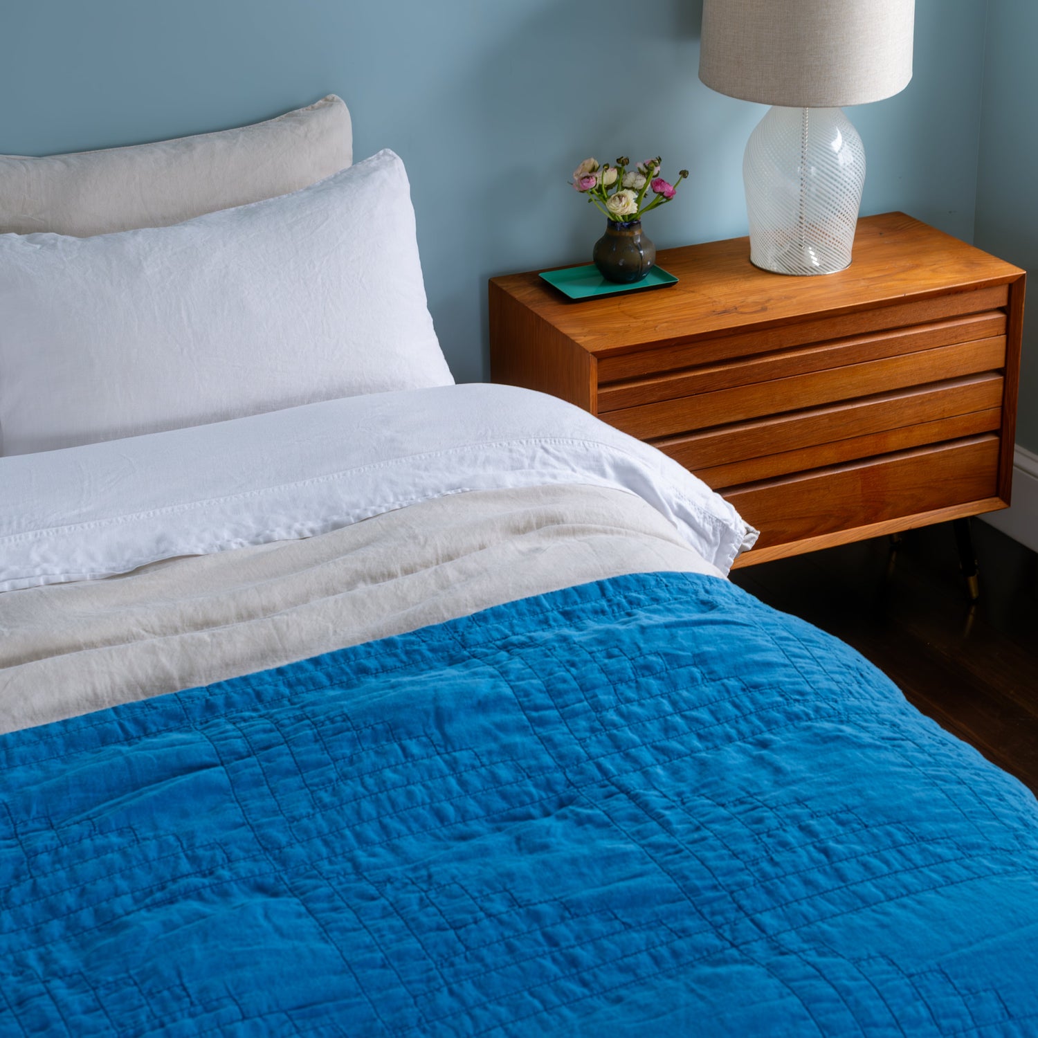 Bedroom with blue quilt, wooden nightstand, lamp, and decorative items.