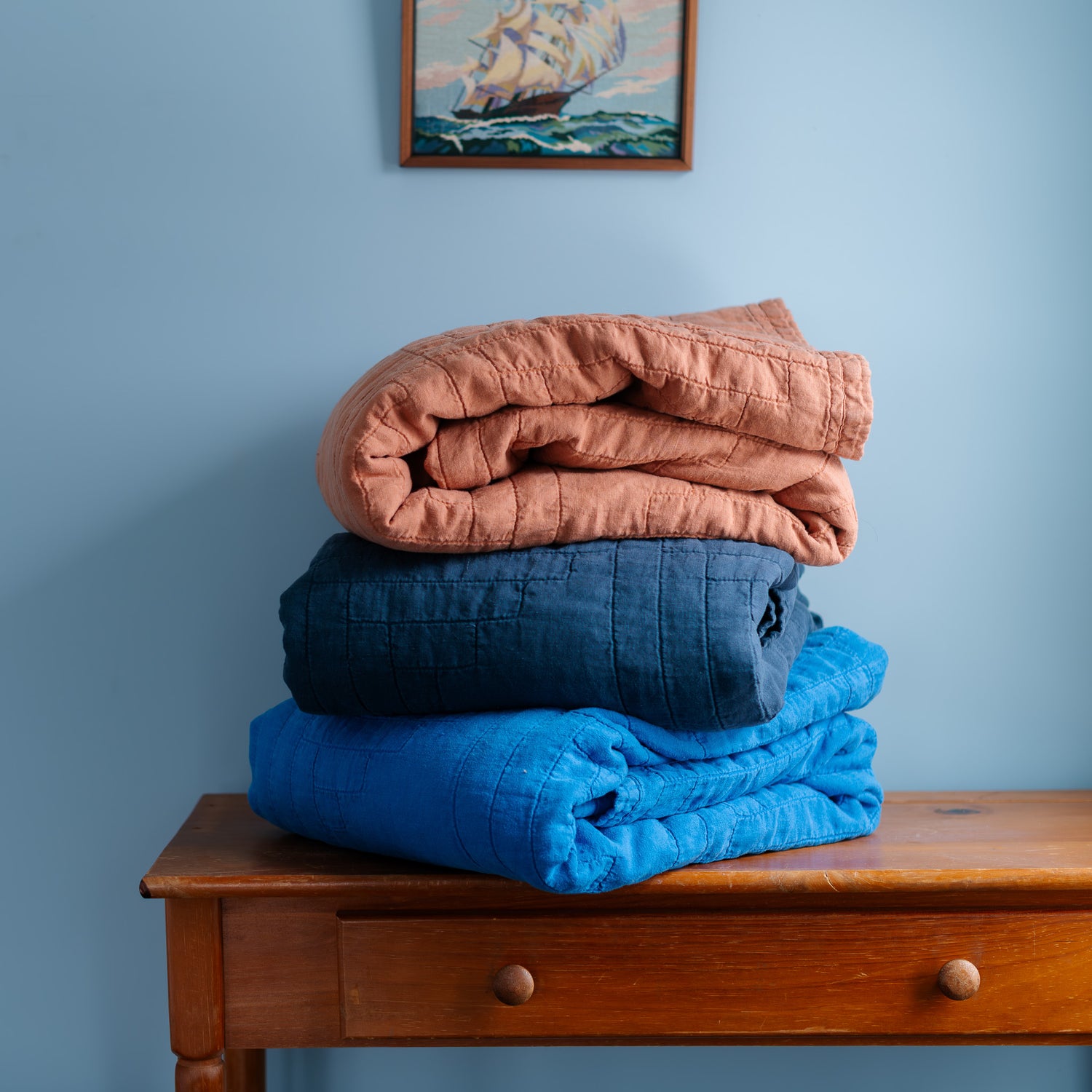 Stack of folded blankets in orange, blue, and navy blue on a wooden bench against a blue wall with a painting.