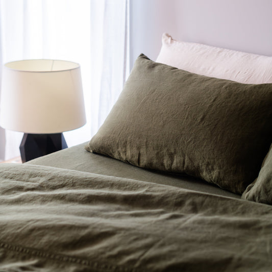 Bed with green sheets and bedding and a lamp in a room with white curtains.