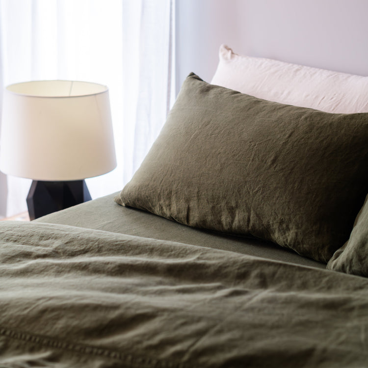 Bed with green sheets and bedding and a lamp in a room with white curtains.