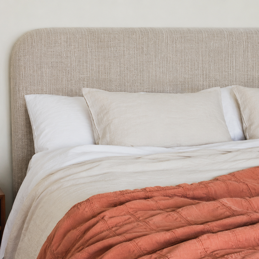 Neatly made bed with beige bedding and a rust-colored throw blanket, nightstand with books and flowers on the side.