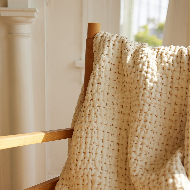 Ivory hand-stiched quilt draped over a wooden chair in a bright room.